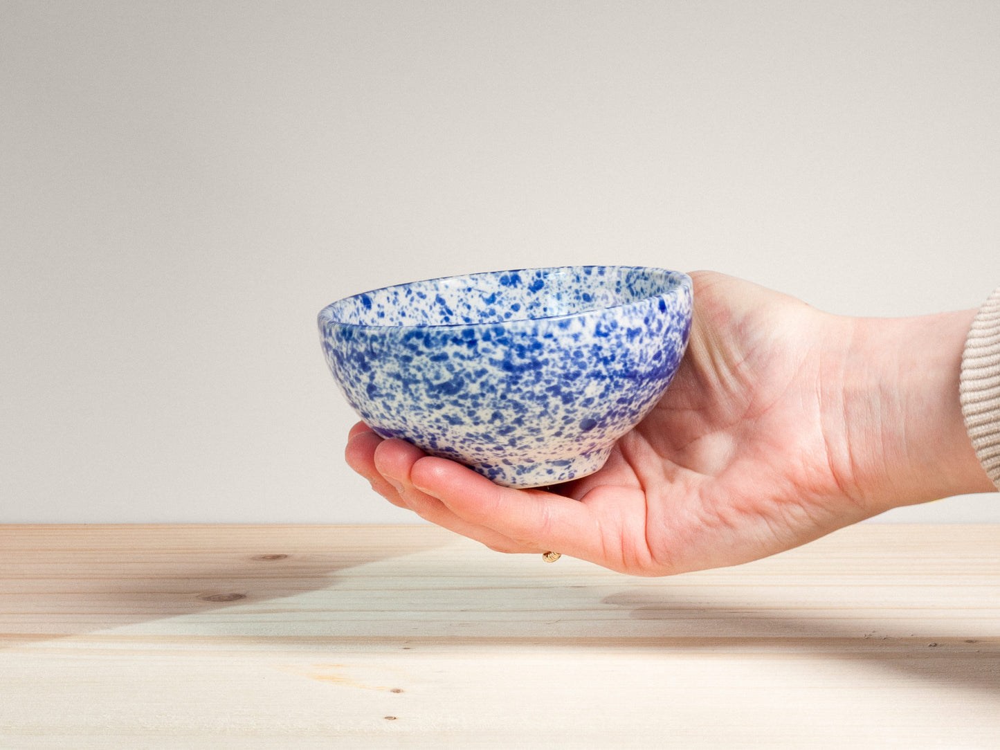 Hand holding a blue and white speckled ceramic bowl on a light wooden surface.