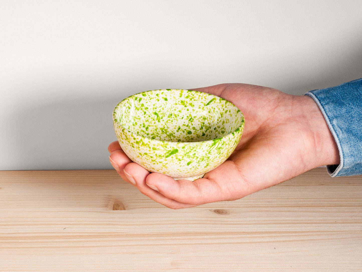 Hand holding a green speckled ceramic bowl on a wooden surface with a light gray background