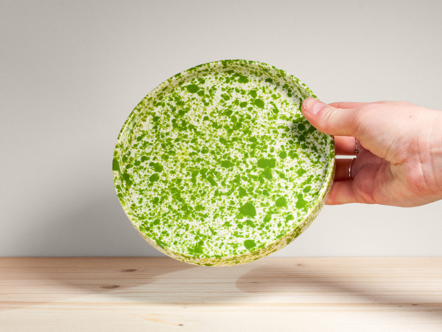 Hand holding a green ceramic plate with white speckles on a wooden surface.