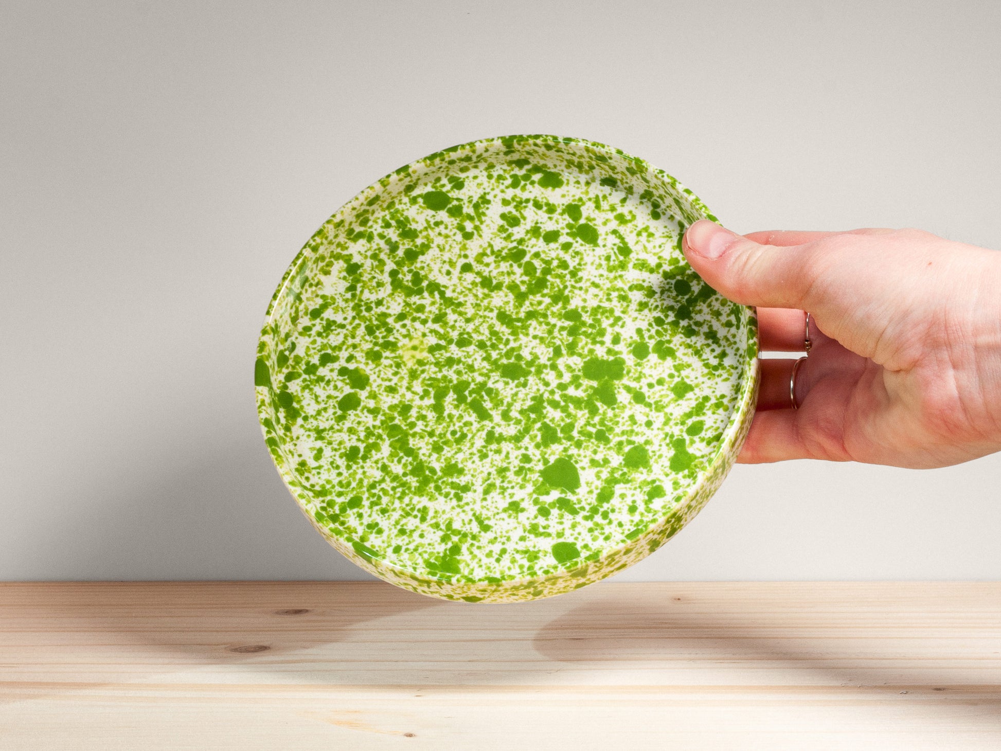 Hand holding a green ceramic plate with white speckles on a wooden surface.