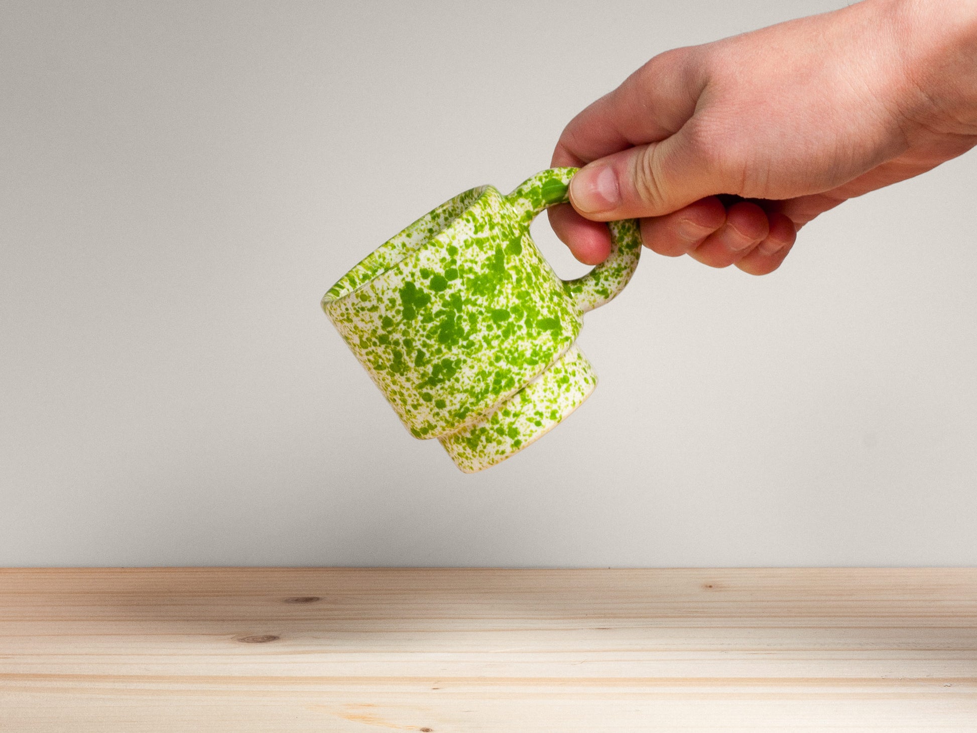 Hand holding a small green and white speckled ceramic mug on a plain background.