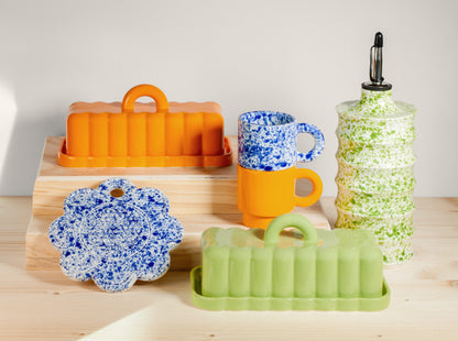 Colorful kitchen items including a green butter dish, orange butter dish, and blue speckled garlic grater on a wooden surface.