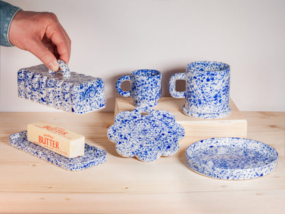 Speckled blue and white ceramic items including mugs, a plate, and a butter dish on a wooden surface.
