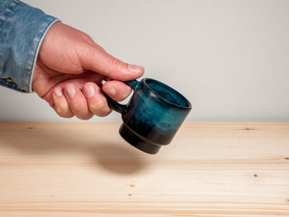 Hand holding dark teal ceramic espresso mug on wooden surface