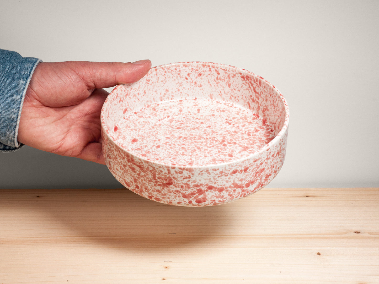 Hand holding a pink and white ceramic bowl on a wooden surface with plain background