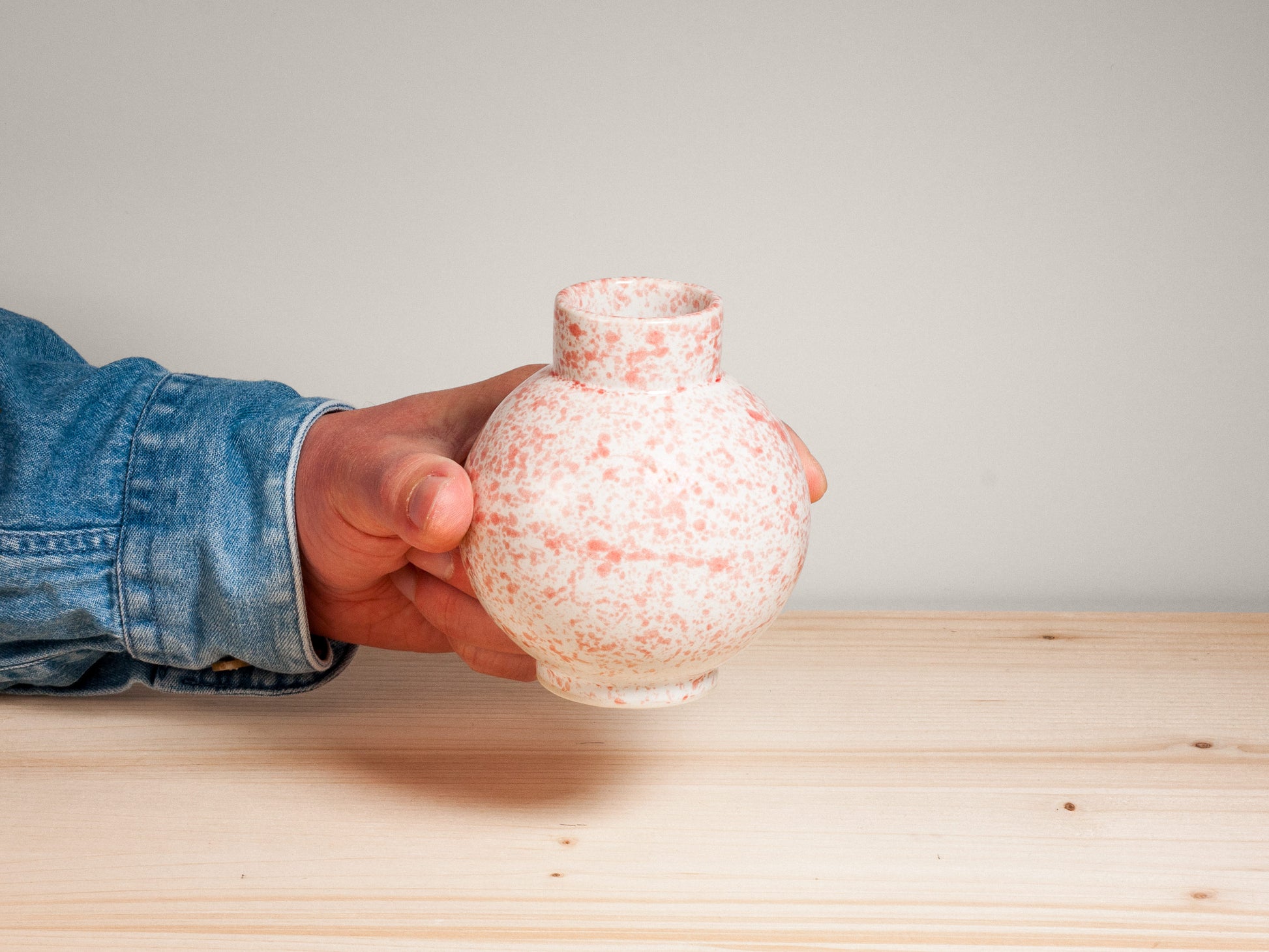 Hand holding a pink and white ceramic vase on a wooden surface with a plain background