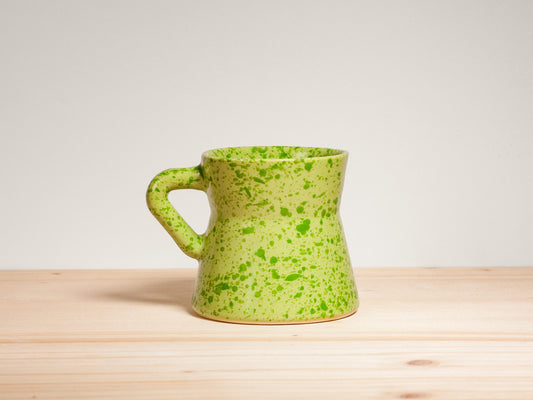 A light green ceramic mug with dark green speckled glaze, featuring a unique tapered shape and a curved handle, set on a light wood surface against a neutral off-white background.