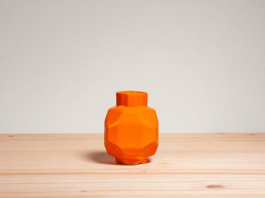 An orange faceted ceramic vase with a matching lid, displayed on a light wood surface against a neutral gray background.