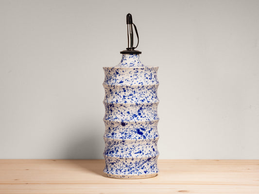 A ceramic oil bottle with a textured, ribbed cylindrical body covered in blue and white speckled glaze, topped with a black metal cap, set on a light wood surface against a neutral gray background.