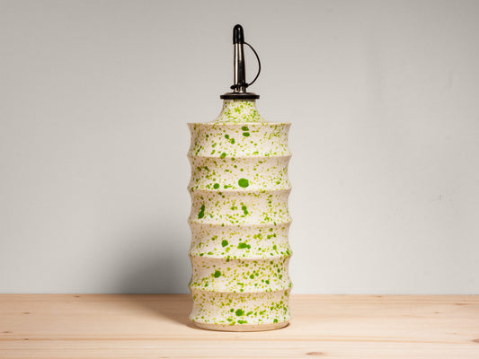 A tall ceramic oil bottle with a ribbed, tiered design, featuring a speckled cream and green glaze pattern. It is topped with a black metal pour spout and is set against a neutral gray background on a light wood surface.