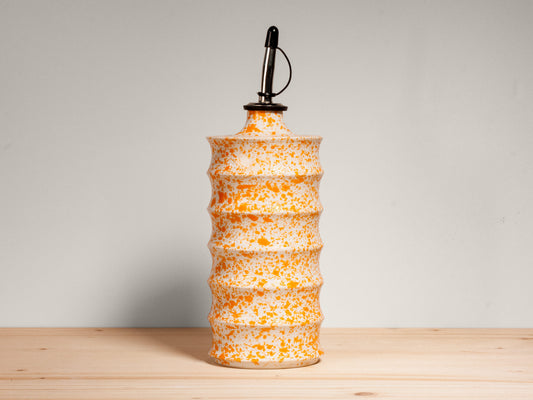 A ceramic oil bottle with a ribbed, tiered body in a speckled orange and white glaze, topped with a black metal oil-dropper cap, set on a light wood surface against a neutral gray background.