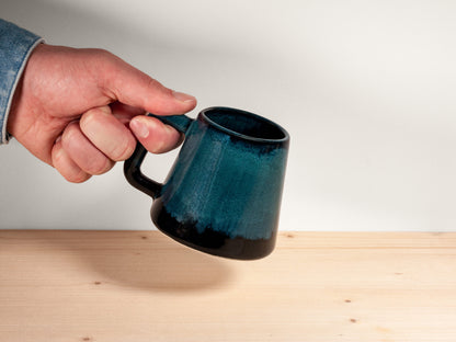 A hand holding a dark teal ceramic mug with a glossy finish and a black base, set against a neutral background with a light wood surface.