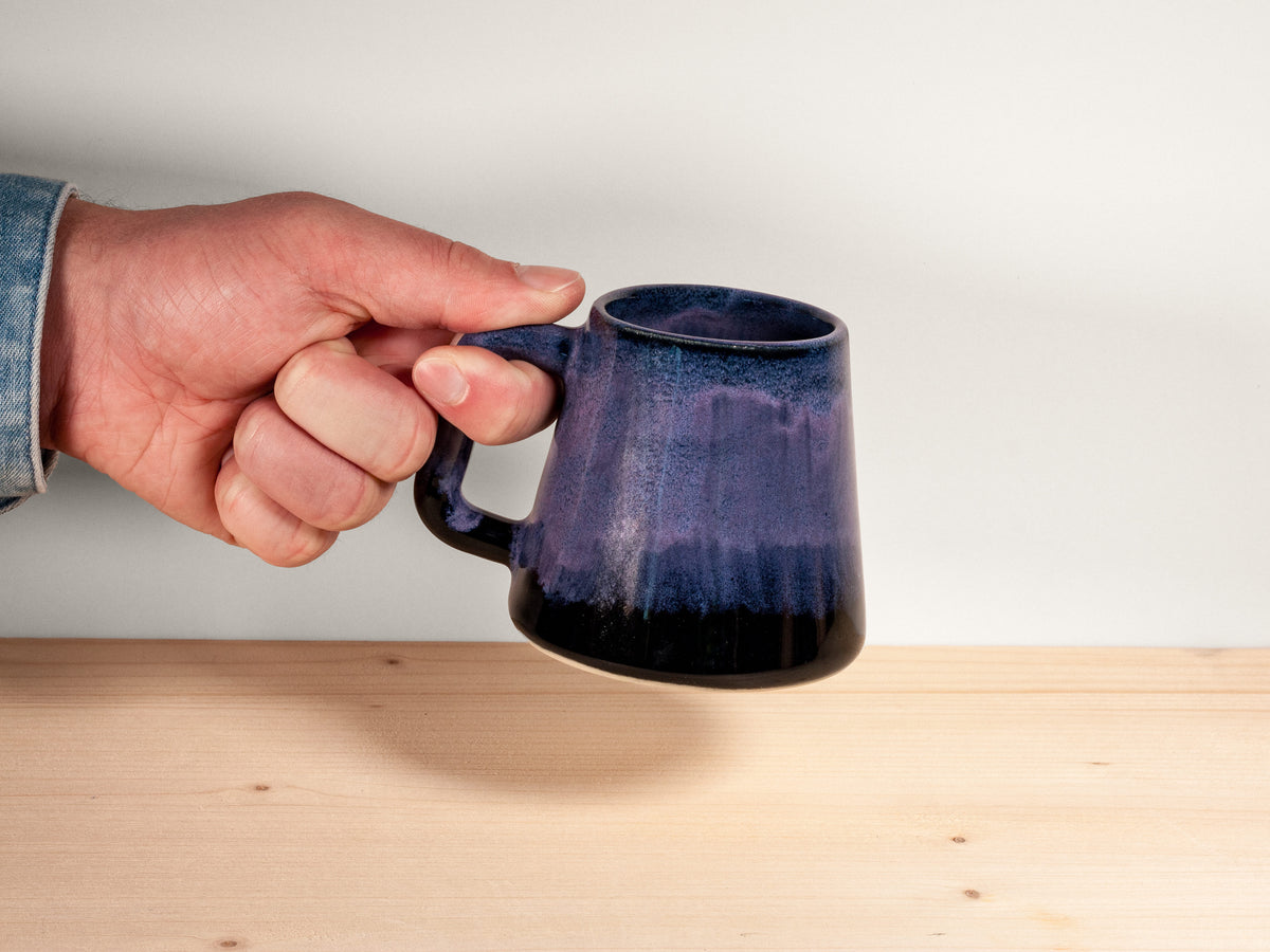 A hand holding a ceramic mug with a dark blue and black gradient glaze, positioned over a light wood surface against a plain white background.