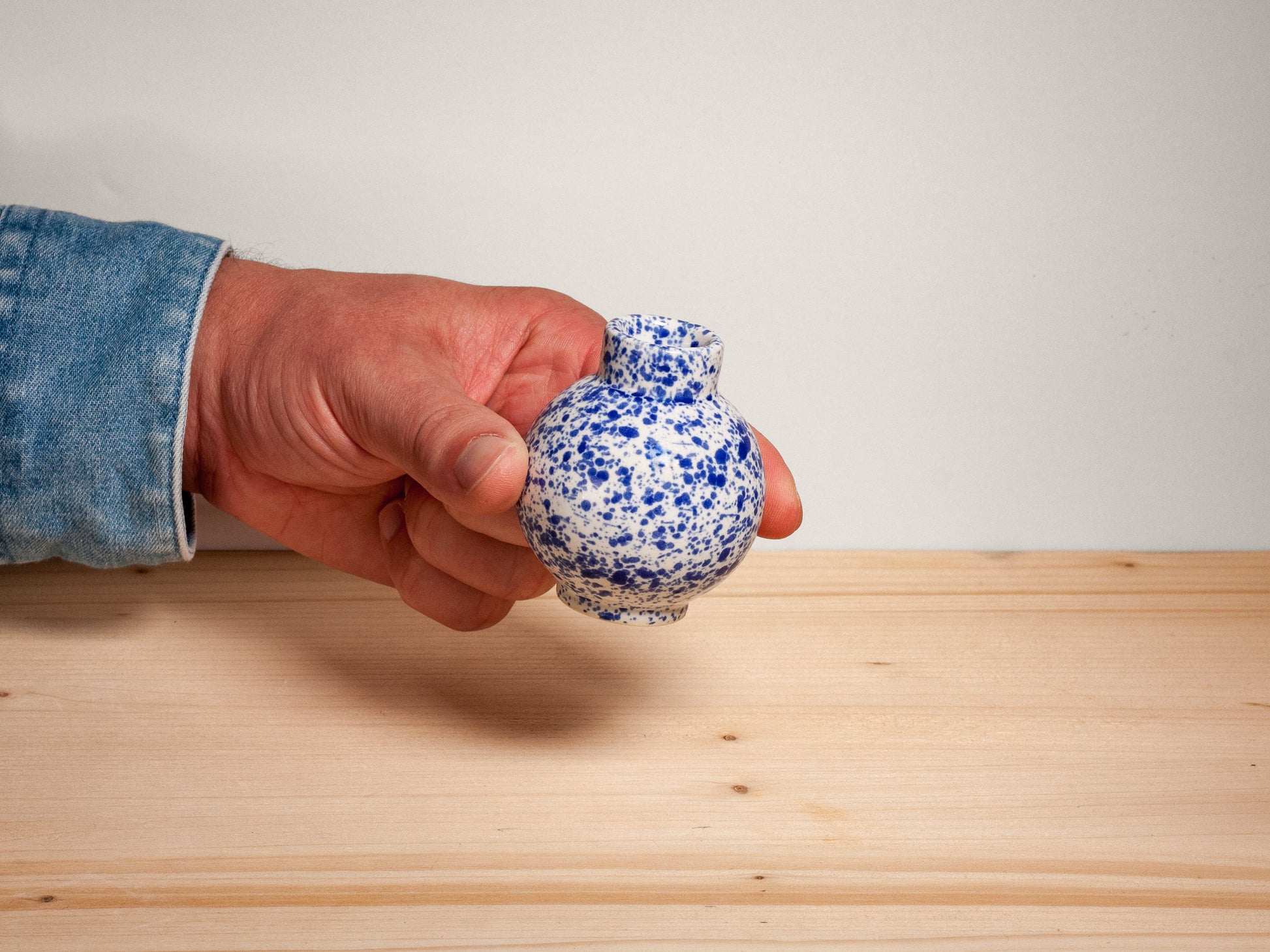 A hand wearing a blue denim sleeve holds a small ceramic vase with a blue and white speckled glaze against a plain light gray wall and a light wood surface.