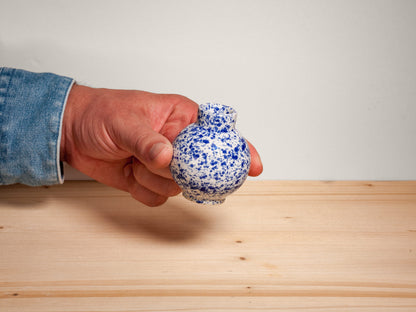 A hand wearing a blue denim sleeve holds a small ceramic vase with a blue and white speckled glaze against a plain light gray wall and a light wood surface.