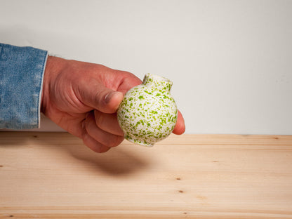 A hand wearing a blue denim sleeve holds a small, rounded ceramic vase with a white glaze and green speckled pattern, positioned over a light wood surface against a plain light gray wall.